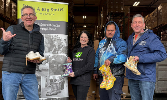 Four people stand beside a Wear A Big Smile Foundation banner in a warehouse, holding up pairs of colorful children's rain boots including unicorn, floral, and animal-print designs.