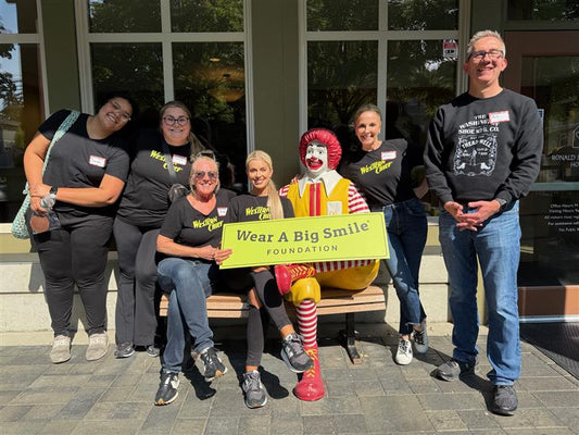 Five people pose with a seated Ronald McDonald statue on a bench, holding a Wear A Big Smile Foundation sign, outside Ronald McDonald House.
