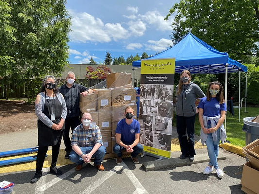 A group of masked volunteers poses outdoors beside a blue Wear A Big Smile Foundation tent and stacked shoe boxes on a hand truck.