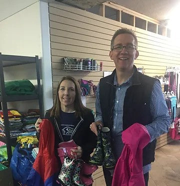 A man and a woman hold up pairs of children's rain boots and raincoats in a shop filled with folded clothing and shoe racks.