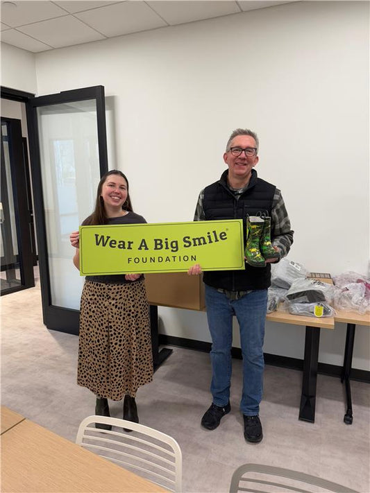 A woman and a man hold a Wear A Big Smile Foundation sign in an office, the man holding a pair of green dinosaur-print rain boots.