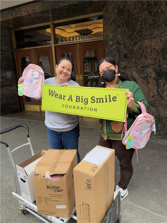 Two women hold a Wear A Big Smile Foundation sign outside a building entrance, each holding a pink backpack, with a luggage cart of boxes in front of them.