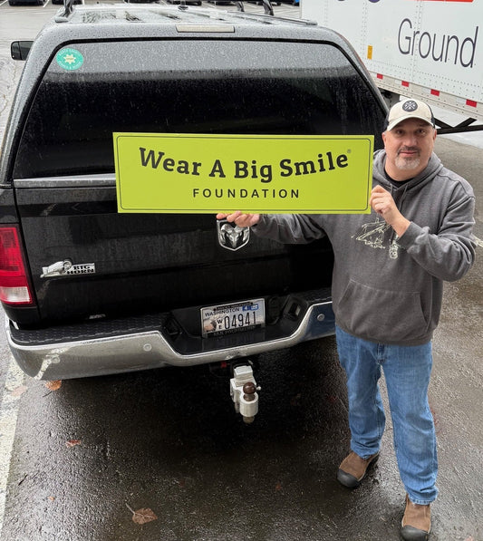 A man stands beside a pickup truck holding a Wear A Big Smile Foundation sign in a parking lot.